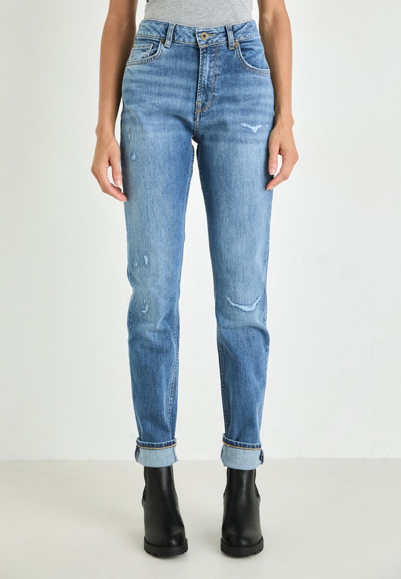 Jeans in denim blu chiaro con vita alta, dettagli usurati e orli arrotolati, abbinati a stivali neri alla caviglia. Texture liscia con dettagli di cucitura.