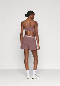 Καφέ αθλητικό σορτς με φαρδιά λευκή μέση που έχει τη φράση "NIKE PRIKE." Συνδυασμένο με αντίστοιχο καφέ αθλητικό σουτιέν, με φαρδιές λουρίδες που διασταυρώνονται στην πλάτη.