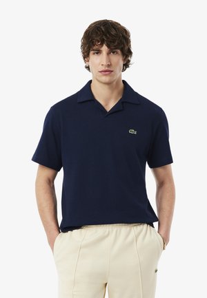 Homme aux cheveux bouclés portant un polo bleu marine à manches courtes et un pantalon crème, les mains dans les poches, sur fond blanc.