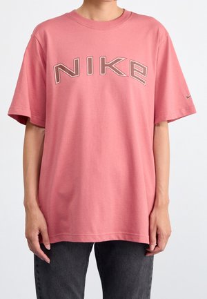 Person iført en løs lyserød Nike t-shirt med stort kontureret logo og lille swoosh på ærmet, parret med mørke jeans.