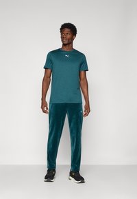 Turkusowa koszulka sportowa z logo Puma, zestawiona z turkusowymi spodniami z weluru. Model ma na sobie czarne buty sportowe z zielonymi akcentami.