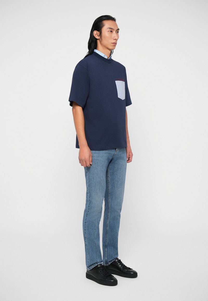 Marineblaues Kurzarmhemd mit einer hellblauen Tasche, kombiniert mit hellblauen Jeans und schwarzen Sneakers. Weicher Stoff; lässiger Schnitt.