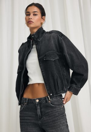 Femme portant une veste en jean noire courte sur un crop top blanc et un jean noir taille haute, debout devant un rideau blanc.
