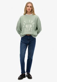 Sudadera oversize verde menta con estampado en el frente, combinada con jeans azules y zapatos negros con cordones. La sudadera tiene hombros caídos y un corte relajado.