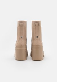 Botines beige con superficie lisa y brillante, laterales elásticos y tacón grueso y cuadrado. Presentan un detalle metálico cerca del tacón.