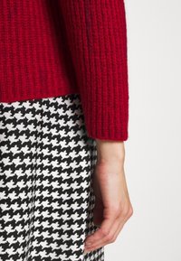 Bras d'une personne portant un pull tricoté rouge à côté d'une jupe à motif pied-de-poule noir et blanc sur un fond clair uni.