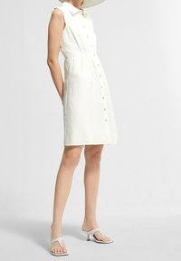 Robe blanche sans manches avec un corsage ajusté et une forme évasée. Tissu lisse avec une texture subtile, associée à des sandales blanches.