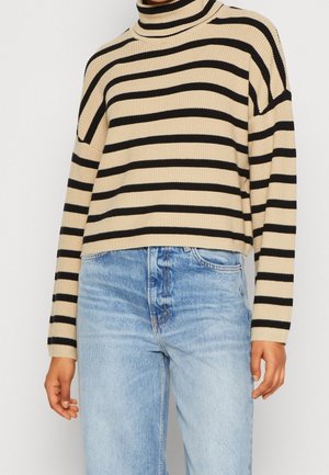 Pull col roulé beige à rayures horizontales noires porté avec un jean taille haute bleu clair sur une personne debout.