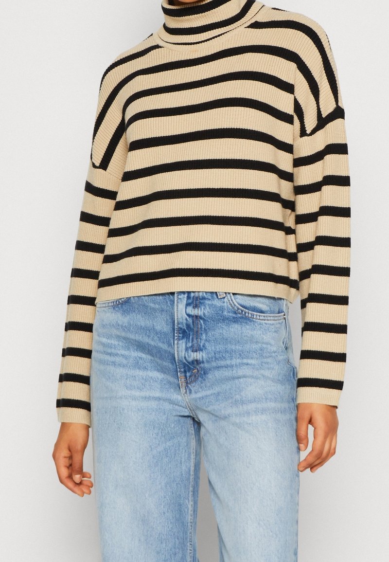 Pull col roulé beige à rayures horizontales noires porté avec un jean taille haute bleu clair sur une personne debout.