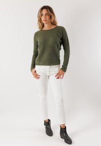 Donna con capelli biondi fino alle spalle che indossa un maglione di lana verde, pantaloni bianchi e stivali neri alla caviglia, in piedi davanti a uno sfondo bianco.
