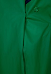 Casaco impermeável verde com textura suave, fecho de botão e um leve caimento. Apresenta um detalhe de gola em azul-marinho contrastante.