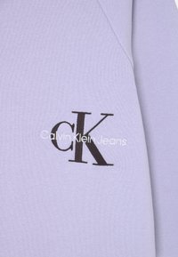 Levandulová bavlna s černým logem "cK" a bílým nápisem "Calvin Klein Jeans" umístěným ve středu. Měkká textura s jednoduchým designem.