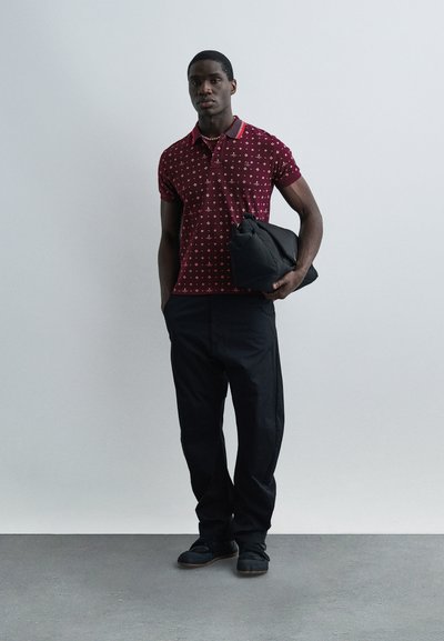 Άνδρας με πουκάμισο πόλο μπορντό με σχέδια και μαύρο παντελόνι κρατάει μια μαύρη τσάντα ρολό μπροστά σε απλό γκρι φόντο.