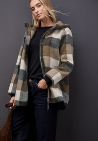 Kostkovaný kabát v šedé, hnědé a krémové barvě, se zvýšenou kapucí, zipovým zapínáním a patentkami. V kombinaci s černým topem a tmavými džínami.