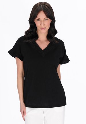 Sötét hullámos hajú nő fekete blúzban, amely csipkés V-nyakkivágással és fodros rövid ujjakkal rendelkezik, fehér háttér előtt áll.