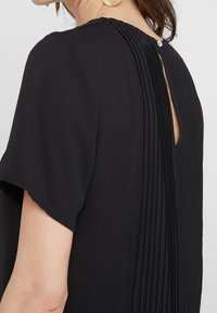 Robe noire avec un dos plissé, manches courtes et une ouverture en forme de goutte d'eau maintenue par un bouton au niveau du col. Texture de tissu lisse.