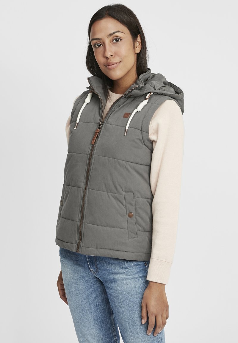 Grau gepolsterte Weste mit Kapuze, ausgestattet mit einem Reißverschluss, Kordelzügen und zwei Seitentaschen mit braunen Akzenten. Getragen über einem leichten Pullover.
