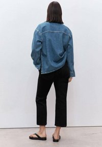 Giacca di jeans azzurra chiara con una vestibilità rilassata, caratterizzata da un orlo curvo e polsini con bottone, abbinata a pantaloni neri a tre quarti e sandali neri.