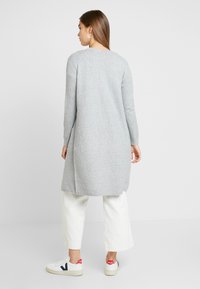 Grauer langer Cardigan mit langen Ärmeln und geradem Schnitt, kombiniert mit cremefarbenen weiten Hosen und weißen Sneakers mit roten und marineblauen Akzenten.