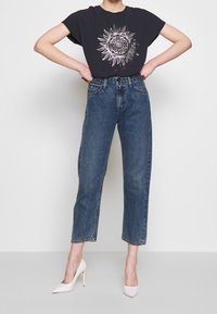 Camiseta negra con un gráfico de sol blanco en el centro, combinada con jeans de denim azul de cintura alta y tacones desnudos. Silueta plana y estructurada.