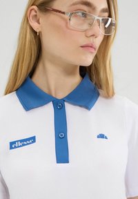 Biała koszulka polo z niebieskim kołnierzykiem i guzikami, z niebieskim logo na piersi i poziomą żebrowaną teksturą na kołnierzyku.