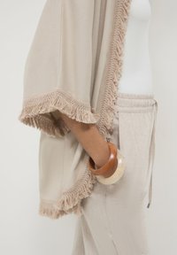 Donna che indossa uno scialle beige con frange, pantaloni beige chiaro con cordino e due bracciali robusti, uno in legno e uno di colore crema, al polso.