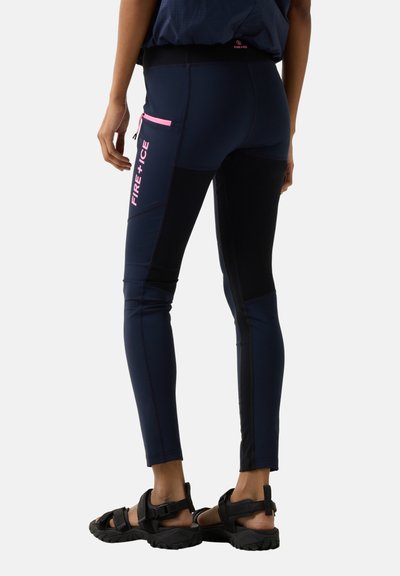 Person iført marineblå og sorte slim-fit bukser med lyserød "FIRE + ICE" tekst på sidelommen, kombineret med sorte sandaler med åbne tæer.