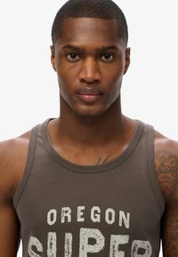 Bruine katoenen tanktop met een ronde hals, voorzien van witte tekst "OREGON SUPER." Zachte textuur met een aansluitende pasvorm.