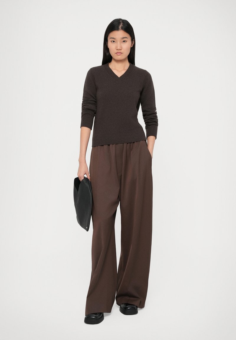Woman wearing a dark brown V-neck sweater and wide-leg brown pants, holding a black clutch bag, standing against a plain background.