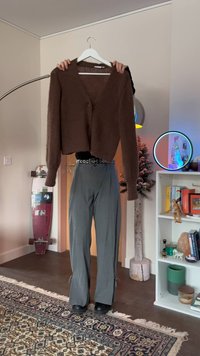 Person iført grå bukser og svarte sko holder en brun cardigan på en hvit kleshenger i en stue med skateboard og bokhylle.