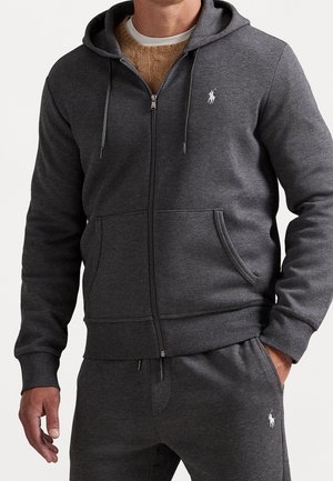 Homme portant un sweat à capuche zippé gris foncé et un pantalon de survêtement assorti avec des logos blancs brodés sur la poitrine et la cuisse.