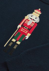 Nėriniuotas svarkelis tamsiai mėlynos spalvos su išsiuvinėtu riešutų trynumo dizainu raudona, žalia ir auksine spalvomis, su sudėtingais uniformos ir batų detalėmis.