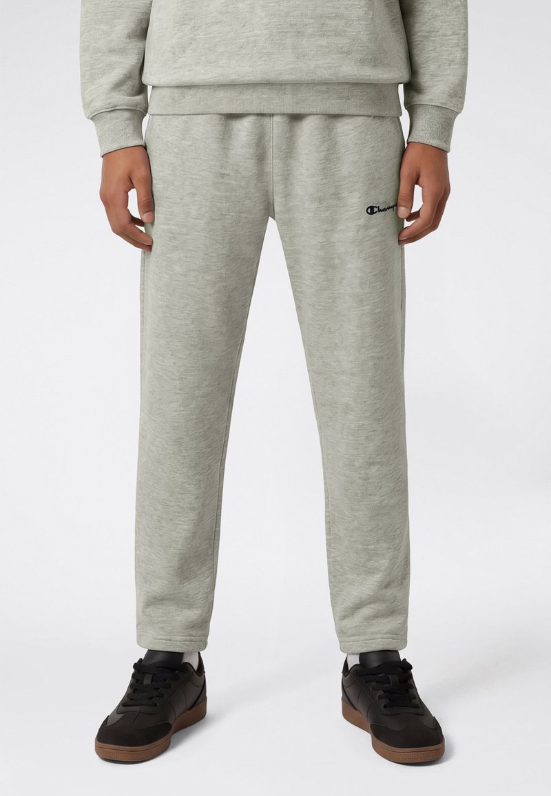 Homme portant un pantalon de jogging gris clair avec le logo Champion sur la cuisse, associé à des baskets noires et un sweat assorti, montré de la taille vers le bas.
