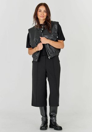 Frau mit langen braunen Haaren, die eine schwarze Lederweste, ein schwarzes, kurzärmliges Oberteil, schwarze knöchellange Hosen und schwarze Stiefel bis zum Knie trägt.