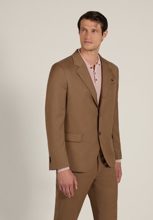Hombre con traje marrón y polo rosa claro, de pie mirando ligeramente de lado sobre un fondo liso.
