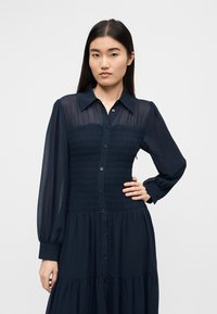 Μακρυμάνικο φόρεμα σε ναυτικό μπλε χρώμα με κουμπιά στο μπροστινό μέρος, μπούστο με σφηκοφωλιά και φουσκωτή φούστα με βολάν, κατασκευασμένο από ελαφρύ ύφασμα.