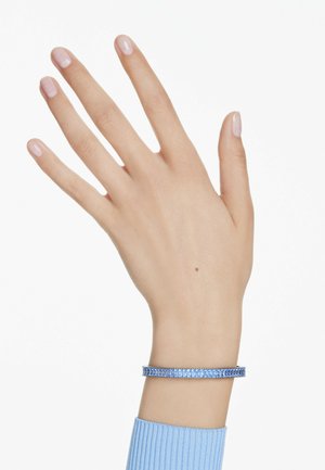 Pulsera de plata con una fila de cristales azules, llevada en una mano de tono de piel claro; puño de tela texturizada con estrías en azul claro.