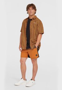 Kortærmet, mønstret skjorte i orange og brune nuancer, parret med orange shorts. Hvide sneakers fuldender outfittet.