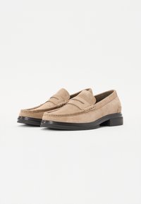 Mocassins en daim beige avec un design cousu, bout rond et un faible talon noir. Texture lisse et doubles coutures décoratives à l'avant.