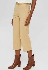 Pantalon beige sur mesure, coupé court, avec des poches latérales et une texture lisse, assorti à des bottines marron à bout carré et talon bloc.