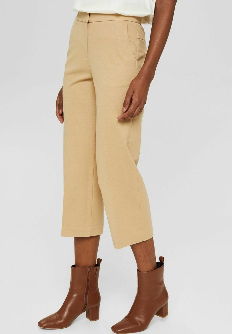 Pantalon beige sur mesure, coupé court, avec des poches latérales et une texture lisse, assorti à des bottines marron à bout carré et talon bloc.