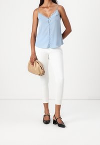 Femme portant un débardeur en dentelle bleu clair, un pantalon slim blanc, des chaussures à talons carrés noires, tenant une pochette douce beige d'une main.