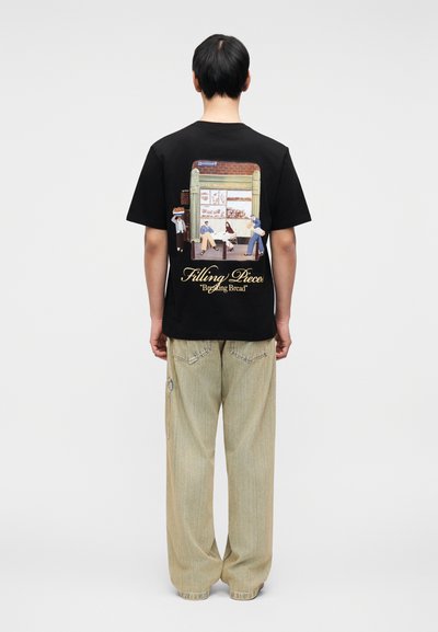 Μαύρο βαμβακερό μπλουζάκι με γραφικό στην πλάτη που απεικονίζει φιγούρες μπροστά σε ένα εστιατόριο. Συνδυασμένο με ανοιχτόχρωμο παντελόνι με φαρδιά παντελόνια.