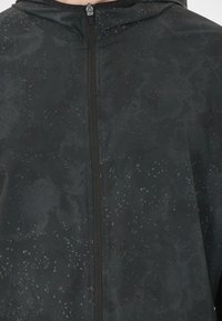 Schwarze wasserfeste Jacke mit einem strukturierten Muster und Reißverschluss, ausgestattet mit einer Kapuze und Branding. Das Material ist glatt mit einer leicht reflektierenden Oberfläche.