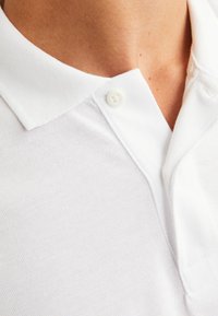 Polo bianco con tessuto texturizzato, colletto a pieghe e un singolo bottone al collo. Design pulito e minimalista.