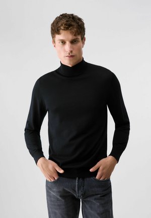 Jeune homme aux cheveux bouclés portant un pull à col roulé noir et un jean foncé, debout les mains dans les poches devant un fond uni.