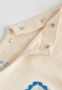 Beige babyromper met metalen drukknoopjes op de schouder en een klein, kleurrijk geborduurd ontwerp vlak bij de onderrand.