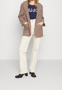 Kvinna som bär en brun hundtandblazer, en marinblå grafisk t-shirt, högmidjade vita byxor och svarta klackskor med fyrkantig tå.