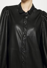 Chemise en cuir noir avec un col à revers, fermeture à boutons sur le devant et manches longues bouffantes. Texture lisse et coupe ajustée.