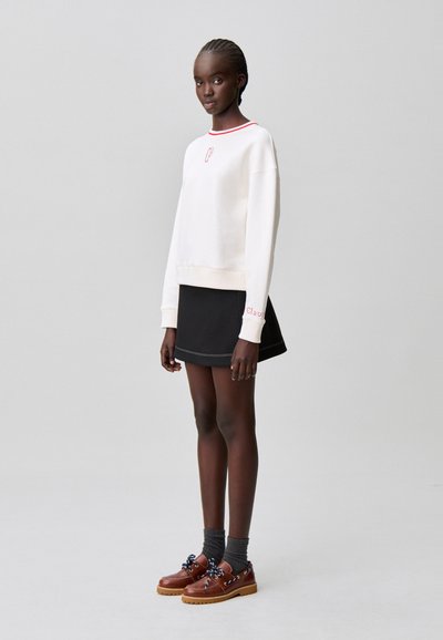 Sweat-shirt de couleur claire avec un col rouge, associé à une jupe noire. Chaussures marron avec des lacets bleu marine et des chaussettes grises. Design simple et texture lisse.